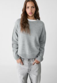 Hellgrauer Pullover mit geripptem Saum und Bündchen, mit rundem Halsausschnitt. Kombiniert mit hochgeschnittenen, hellgrauen Jeans.