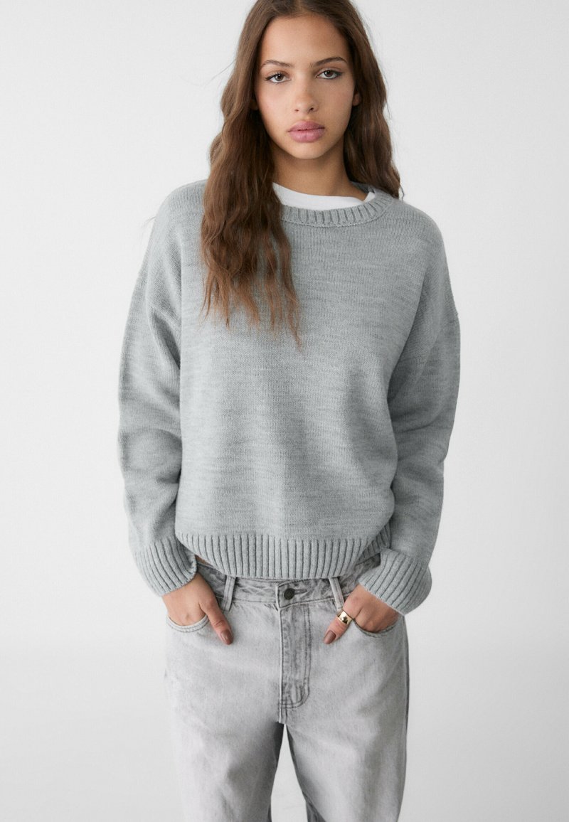 Hellgrauer Pullover mit geripptem Saum und Bündchen, mit rundem Halsausschnitt. Kombiniert mit hochgeschnittenen, hellgrauen Jeans.