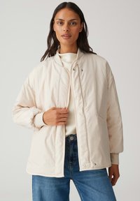 Femme portant une veste matelassée beige clair sur un pull crème et un jean bleu, ajustant l'avant de sa veste avec sa main.