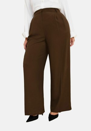 Kobieta ubrana w brązowe, szerokie spodnie z wysokim stanem, białą bluzkę i czarne, spiczaste szpilki, stojąca na jednolitym tle.