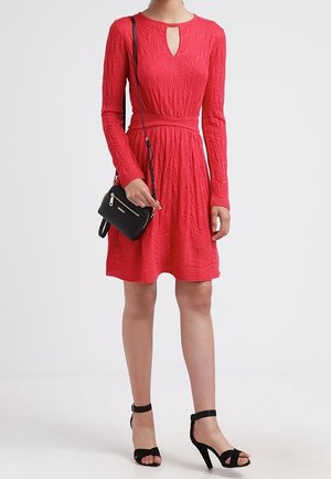 Femme portant une robe rouge texturée à manches longues et longueur genoux, des sandales à talons aiguilles noires, et tenant un petit sac bandoulière noir.