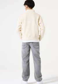 Sudadera color crema con cuello alto, combinada con jeans sueltos de color gris claro con costuras en los bolsillos traseros y zapatillas blancas.