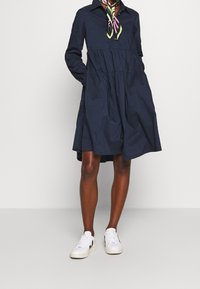 Marinblå klänning med krage och åtsittande byst samt volangkjol. Har långa ärmar och en slät textur. Bärs med vita sneakers.