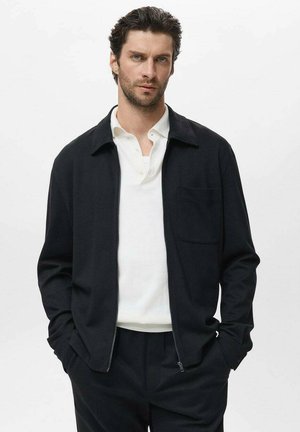 Chaqueta negra con cremallera y cuello tipo camisa, con un bolsillo en el pecho, llevada sobre una camiseta polo blanca; tela suave con un corte entallado.