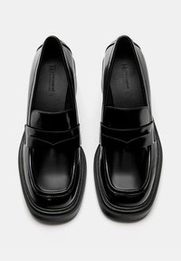Ein Paar schwarze glänzende Lederloafers mit quadratischen Zehen und dicken Sohlen, von oben auf einem weißen Hintergrund betrachtet.