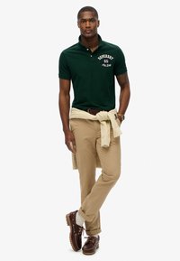 Polo shirt vert foncé avec logo blanc, pantalon kaki et chaussures bateau marron. Pull noué autour de la taille, manches retroussées et montre visible.
