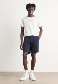 Hvitt bomull t-skjorte med trykt logo, kombinert med marineblå shorts og hvite sneakers. Enkel design med korte ermer og avslappet passform.