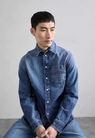 Camicia button-up in denim con tonalità di blu chiaro e scuro, maniche lunghe, tasca sul petto con il logo "VERSACE" ricamato e colletto classico.