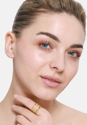 Goldring mit drei gestapelten Bändern, die strukturierte und glatte Oberflächen aufweisen. Die Hand des Models ruht nahe dem Kinn vor einem neutralen Hintergrund.