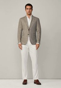 Blazer gris clair avec revers, deux boutons et détails de poche, porté sur une chemise blanche et associé à un pantalon blanc et des chaussures marron.