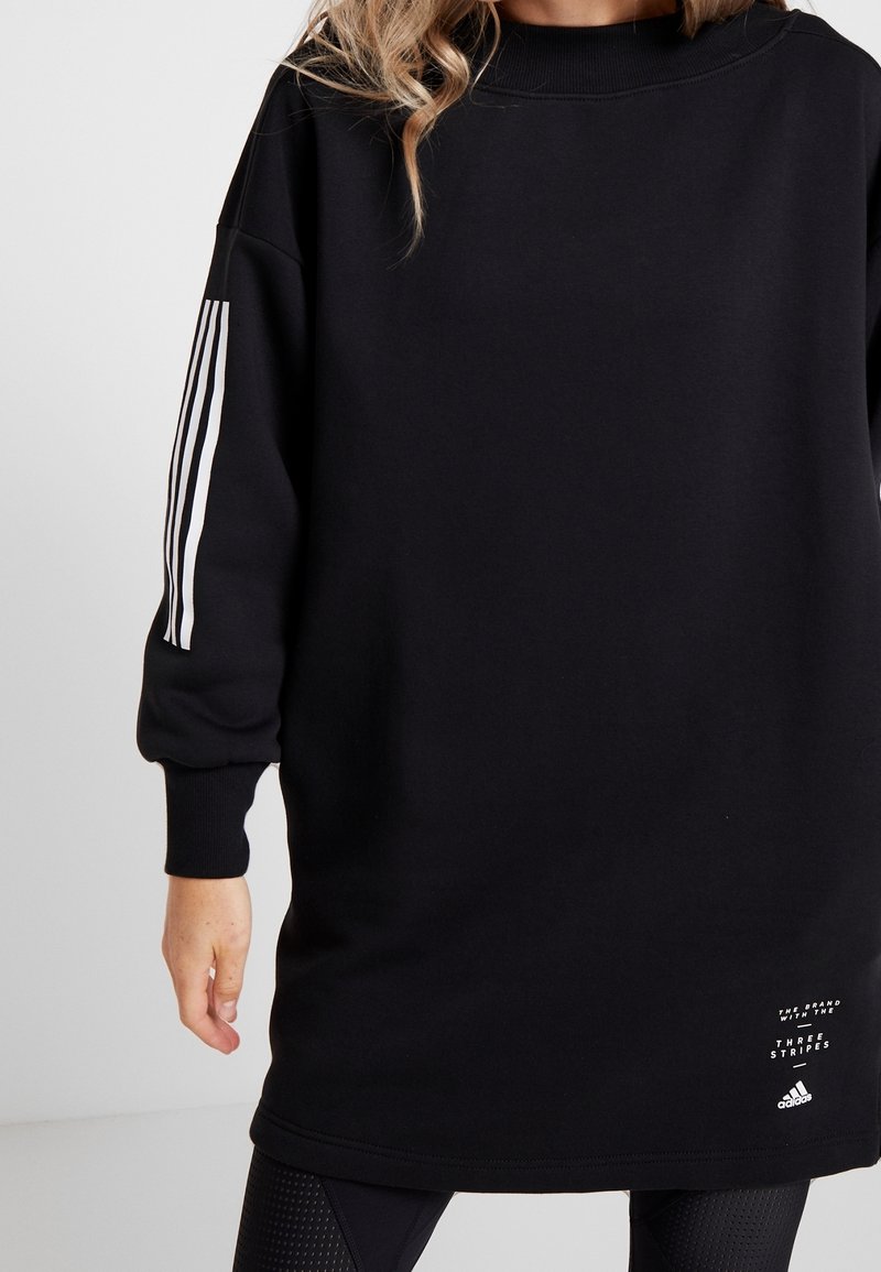 Personne portant une robe sweat Adidas noire avec trois bandes blanches sur la manche et un texte de logo près de l'ourlet.