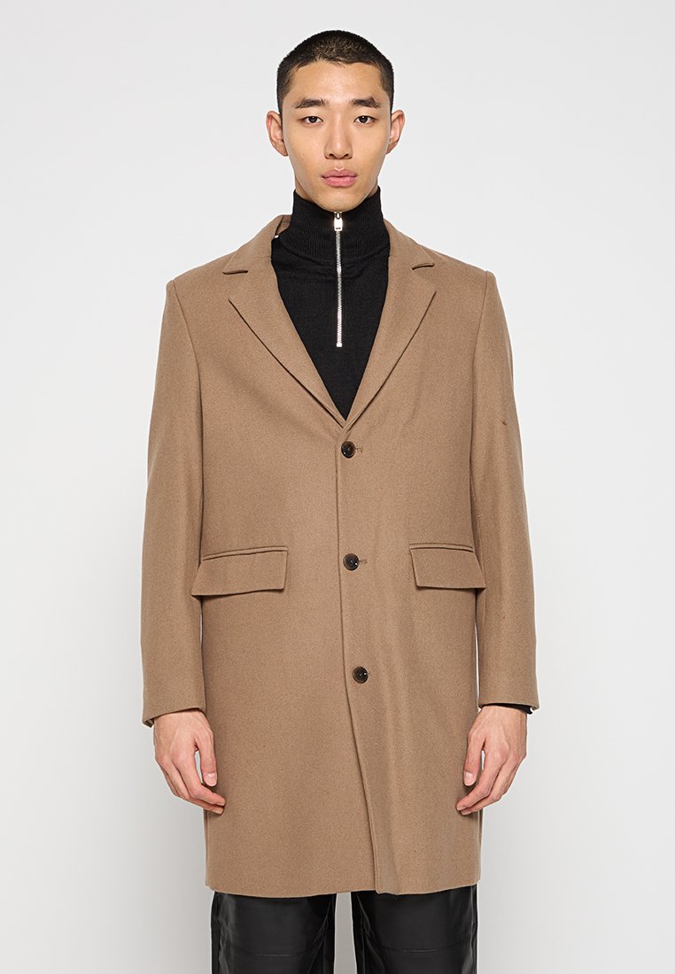 Cappotto in misto lana marrone con design sartoriale, colletto a punta, due tasche frontali e chiusura a bottoni. Indossato sopra un dolcevita nero con zip.