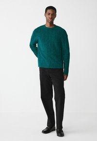 Tuumel roheline kudum, millel on tekstureeritud muster, ümar kaelus, allapoole vajunud õlad, koos mustade pükste ja mustade kingadega.