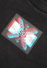 Tessuto nero con un'etichetta rettangolare che presenta elementi grafici rossi e blu, testo e frecce. L'etichetta è cucita sul capo.