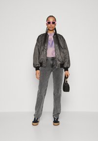Giubbotto bomber nero con finitura testurizzata, top trasparente viola, jeans grigi e sneaker nere con suole in gomma, accessoriate con una piccola borsa nera.