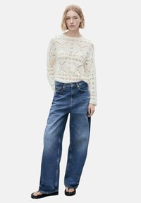 Sweter w kolorze kremowym, robiony na drutach, z wyraźnym wzorem, w połączeniu z luźnymi niebieskimi dżinsami. Modelka nosi czarne klapki.
