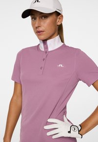 Golf-Polohemd in Lavendel, mit kurzen Ärmeln und einem Knopfkragen. Verfügt über ein weißes Logo auf der Brust und einen weißen Handschuh mit schwarzen Akzenten.