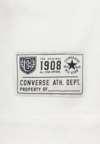 Etiqueta de tela blanca con texto azul marino, que presenta el logo "CONVERSE", "1908" y "DEP. DE ATLETISMO" en una forma rectangular con bordes redondeados.