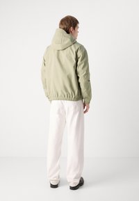 Green hooded jacket with a relaxed fit, elastic cuffs, and waistband, paired with white pants. Smooth texture and minimal design.