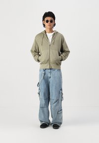 Olivgrüner Reißverschluss-Hoodie, weißes Rundhals-T-Shirt, übergroße blaue Cargo-Jeans mit Taschen, schwarze Schuhe und runde Sonnenbrillen.