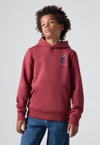 Sudadera con capucha color burdeos, con un bolsillo tipo canguro, puños acanalados y un diseño gráfico en el pecho. Combinada con jeans de mezclilla azules.