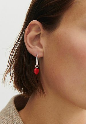 Srebrna kolczyk w kształcie koła z małym, czerwonym serduszkiem noszony na lewym uchu osoby na neutralnym tle.