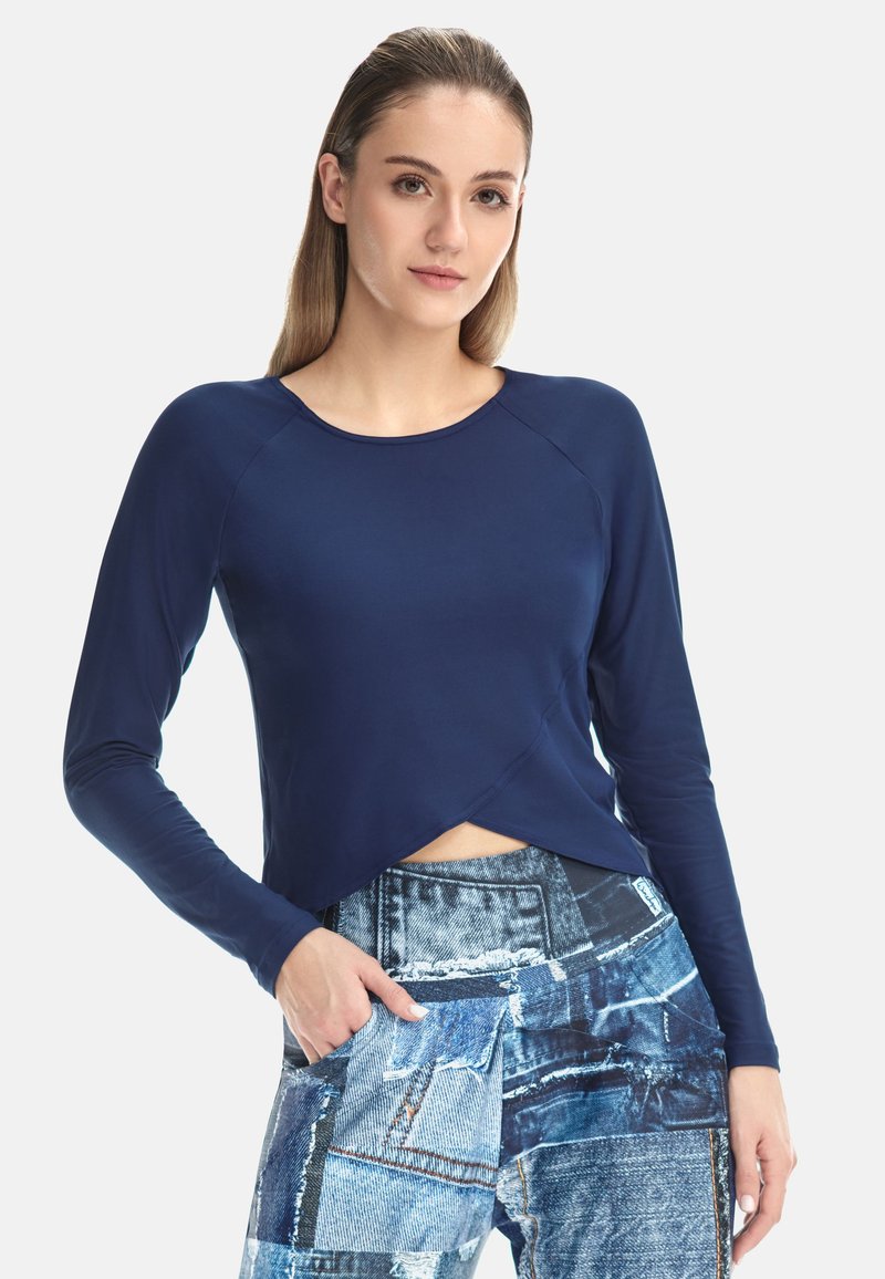 Žena sa ravnom svijetlosmeđom kosom koja nosi tamnoplavu kratku majicu dugih rukava i traperice visokog struka s našicima, stoji s jednom rukom u džepu.