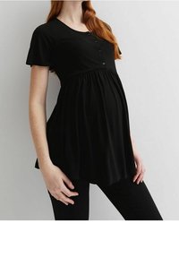 Těhotná osoba v černém těhotenském topu, jemně se dotýká břicha, stojící na jednoduchém světlém pozadí.