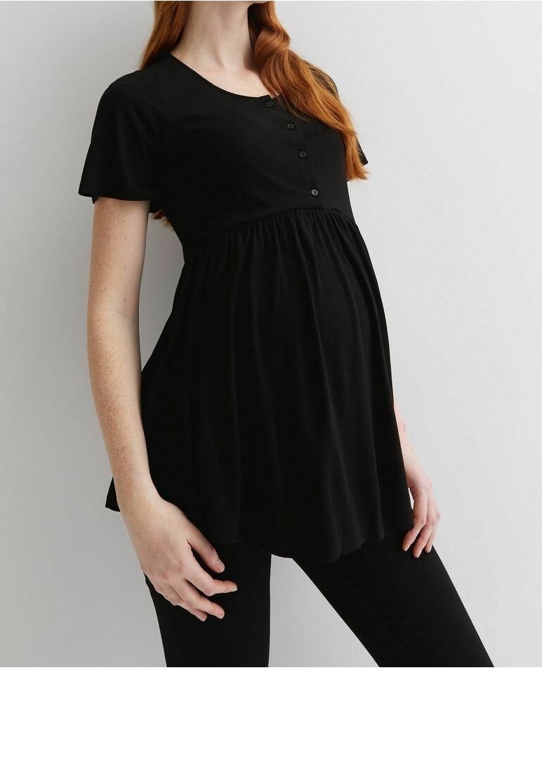 Těhotná osoba v černém těhotenském topu, jemně se dotýká břicha, stojící na jednoduchém světlém pozadí.