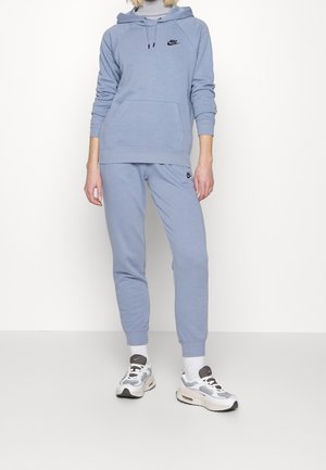 Personne portant un sweat à capuche Nike bleu clair et un jogging assorti avec des baskets et des chaussettes blanches Nike, debout sur un fond uni.