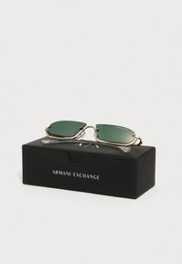 Ochelari de soare cu lentile verzi, ramă din metal aurie și vârfuri transparente, prezentați pe o cutie neagră Armani Exchange cu logo.