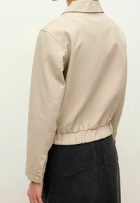 Veste en coton beige avec un col, une taille élastique et des poignets boutonnés. Le design présente une coupe décontractée et un style minimaliste.