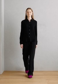 Zwarte lange mouwen blouse met een kraag, gecombineerd met zwarte broek en felle roze schoenen, op een model dat tegen een lichte achtergrond loopt.
