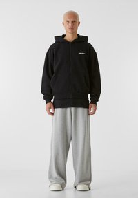 Svart zip-up hoodie med stora fickor, med en logotyp på bröstet. Matchad med ljusgrå, löst sittande sweatpants och vita skor.