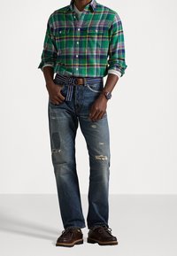 Camicia a quadri verde con bottoni e dettagli bianchi, indossata sopra jeans blu consumati con toppe. Scarpe marrone scuro e cintura a righe completano il look.