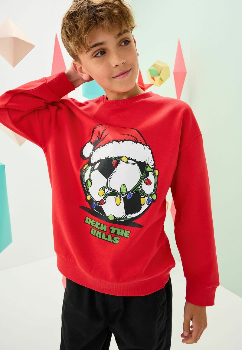 Sweat-shirt rouge avec un graphique de ballon de football enveloppé de guirlandes lumineuses de Noël et d'un chapeau de Père Noël. Le texte dit "DÉCOREZ LES BALLONS". Texture lisse, manches longues.
