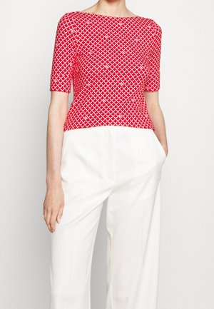 Kobieta ubrana w czerwony krótki top z białymi geometrycznymi wzorami oraz wysokim białymi spodniami, stoi z ręką w kieszeni.