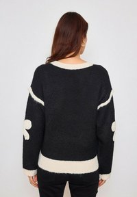 Pull en tricot noir avec des bordures crème et des accents floraux blancs décoratifs sur les manches. Design surdimensionné avec une coupe décontractée.