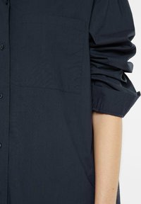Dark blue, long-sleeve shirt made of lightweight fabric. Features a chest pocket, button closure, and gathered sleeve detail.