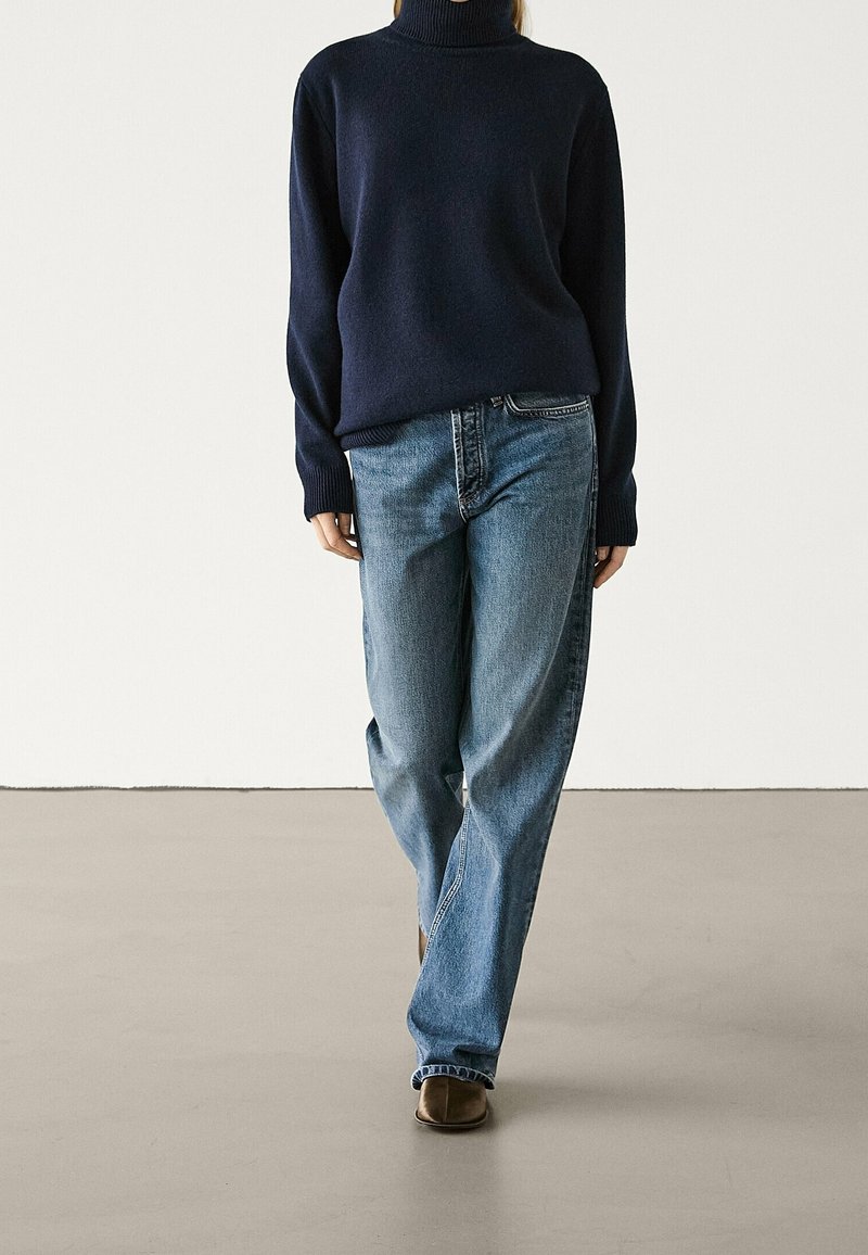 Person wearing a dark navy turtleneck sweater, blue straight-leg jeans, and brown shoes standing on a plain floor against a white wall.