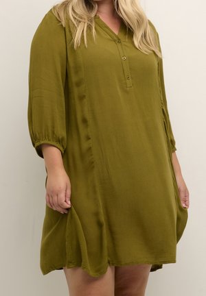 Woman with blonde hair wearing loose olive green long-sleeve dress with buttons, standing against a plain light background.