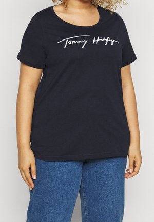 Personne portant un t-shirt Tommy Hilfiger bleu marine et un jean bleu, visible du cou jusqu'aux hanches sur un fond uni.