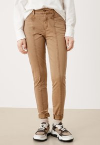 Person, der beiger, schmal geschnittener Hose mit Nahtdetails, weißem Hemd, beigen Socken und beigen und schwarzen klobigen Turnschuhen steht auf einem beigen Boden.