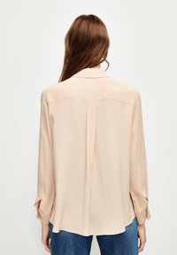 Blusa a maniche lunghe rosa chiaro con colletto, texture liscia e piega sul retro, abbinata a jeans blu, con polsini delle maniche abbottonati.