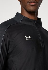 Μαύρο αθλητικό πουκάμισο με φερμουάρ και λεπτό γιακά, κατασκευασμένο από απαλό ύφασμα. Διαθέτει λευκό λογότυπο Under Armour στο στήθος και υφασμάτινες επενδύσεις στους ώμους.