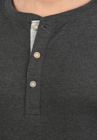 Dunkelgraues Henley-Shirt mit rundem Halsausschnitt, drei Holzknöpfen auf der Vorderseite und einer hellgrauen Innenleiste. Weicher Stoff.