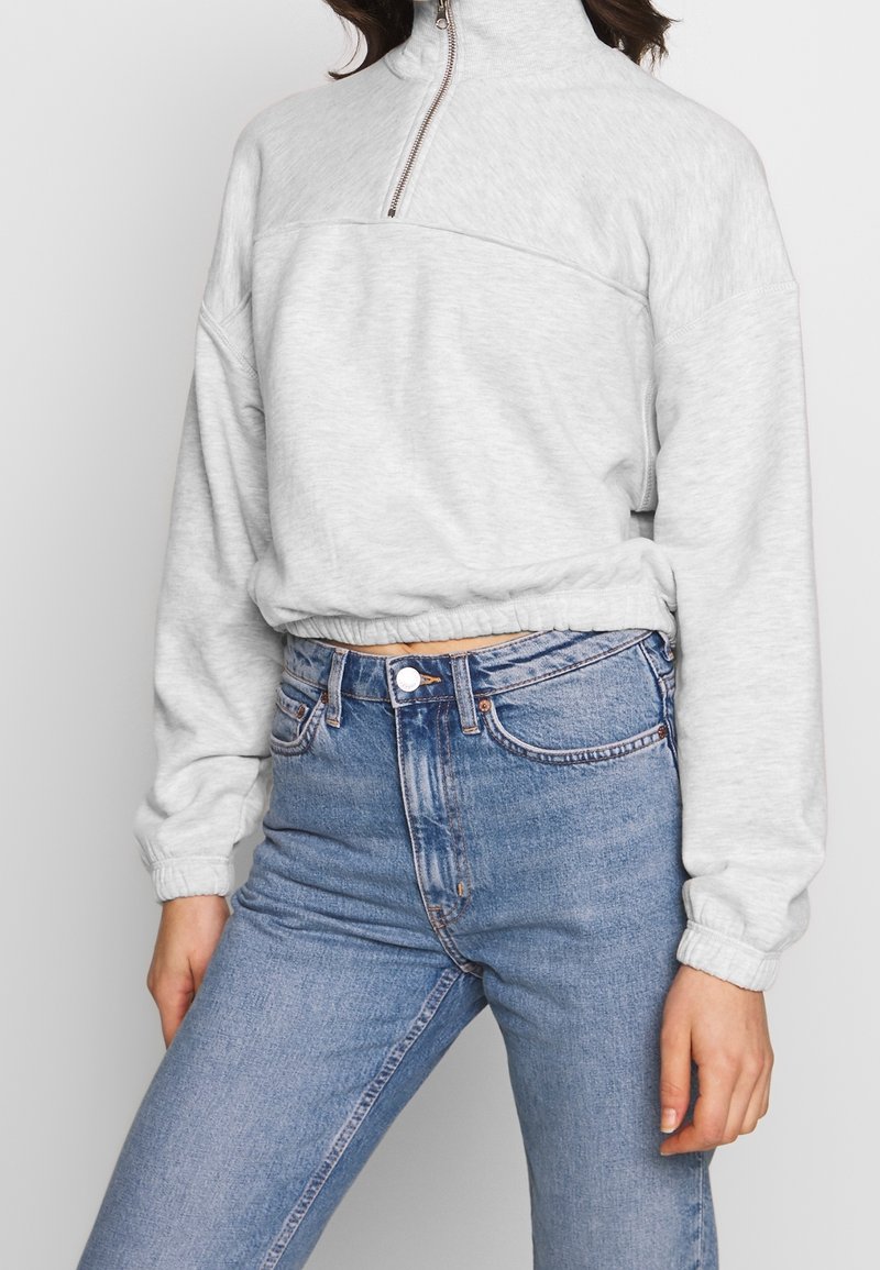 Lichtgrijs sweatjack met rits en hoge kraag, voorzien van lange mouwen en een cropped zoom, gecombineerd met hooggesneden blauwe jeans.