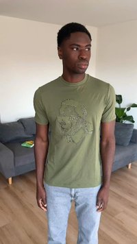 Homme aux cheveux courts et bouclés portant un t-shirt graphique vert olive et un jean bleu clair, debout dans un salon avec un canapé gris et une plante.