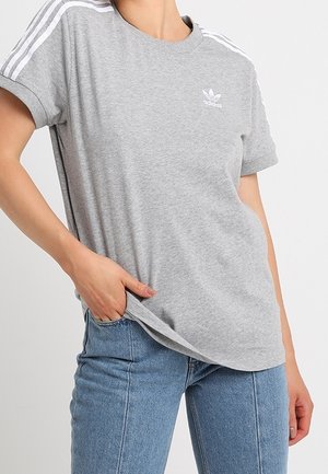 Person iført en grå Adidas t-shirt med hvide striber på ærmerne og logo på brystet, kombineret med lyseblå jeans, hånd i lommen.