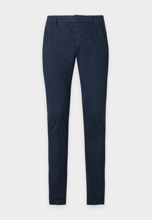 Ein junger Mann steht mit einer Hand in der Tasche, trägt eine hellbeige geknöpfte Jacke über einem olivgrünen T-Shirt, schwarze Hose und off-white Sneaker.